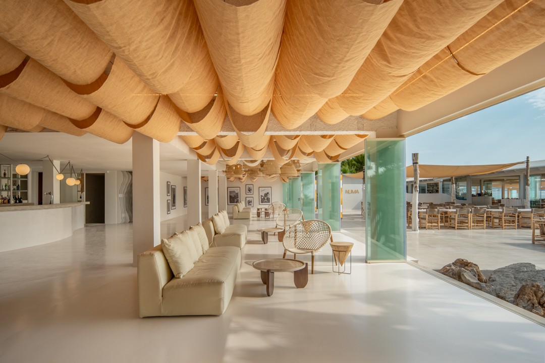 Numa Beach Interior de un restaurante mediterráneo con diseño minimalista, sofás claros, techos decorados con telas onduladas y vista abierta hacia una terraza exterior junto al mar